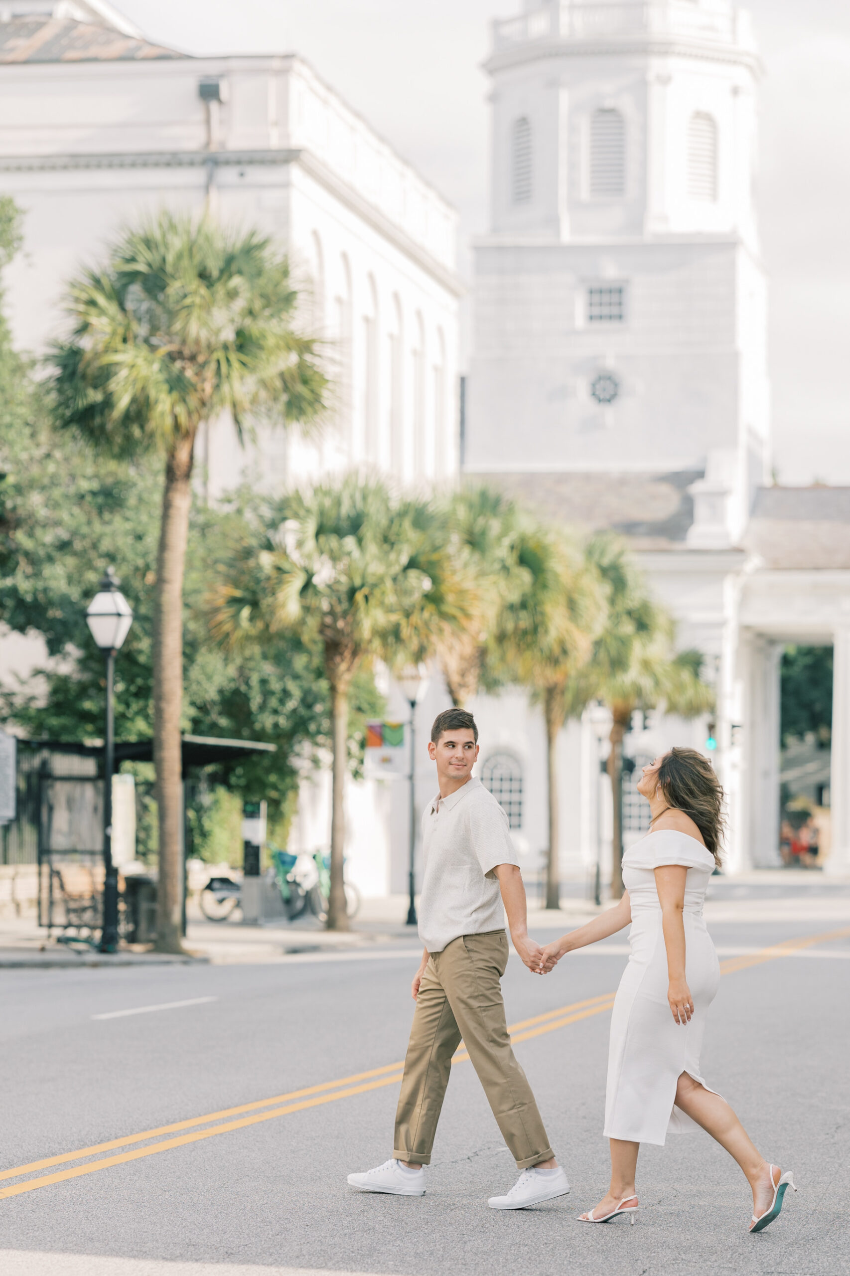 Columbia SC Engagement Photos | Stee Garman Photography Fine-art Wedding Photographer | A couple walks hand in hand across a sunny street, surrounded by palm trees and historic white buildings, conveying a relaxed and romantic mood.