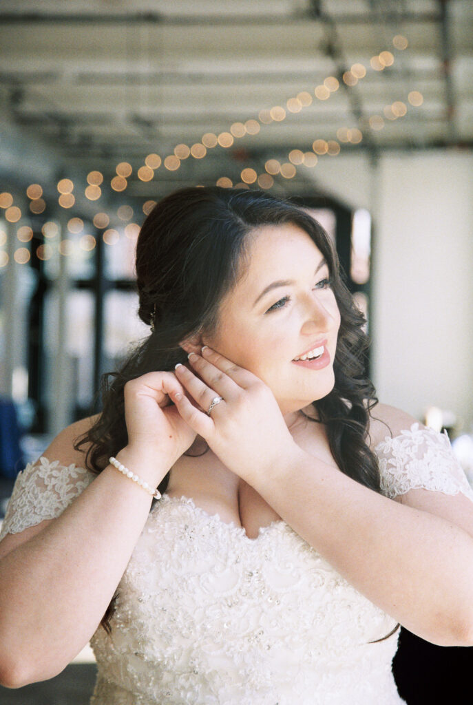 Film Photographer | Bride smiling and looking out the window while putting her earrings in. 35mm color film photographed before wedding. Wedding photos at 701 Whaley in Columbia, South Carolina.