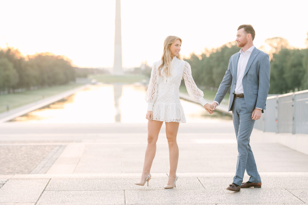 DC Engagement Photo | Couple holding hands and walking in front of the Lincoln Memorial Steps with the Washington Memorial in the background at Sunrise