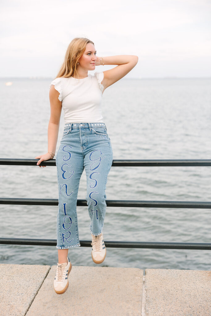 Charleston Senior Pictures | Senior Session in Charleston SC | Senior wearing a white top and custom blue jeans, leans on railing in front of waterfront