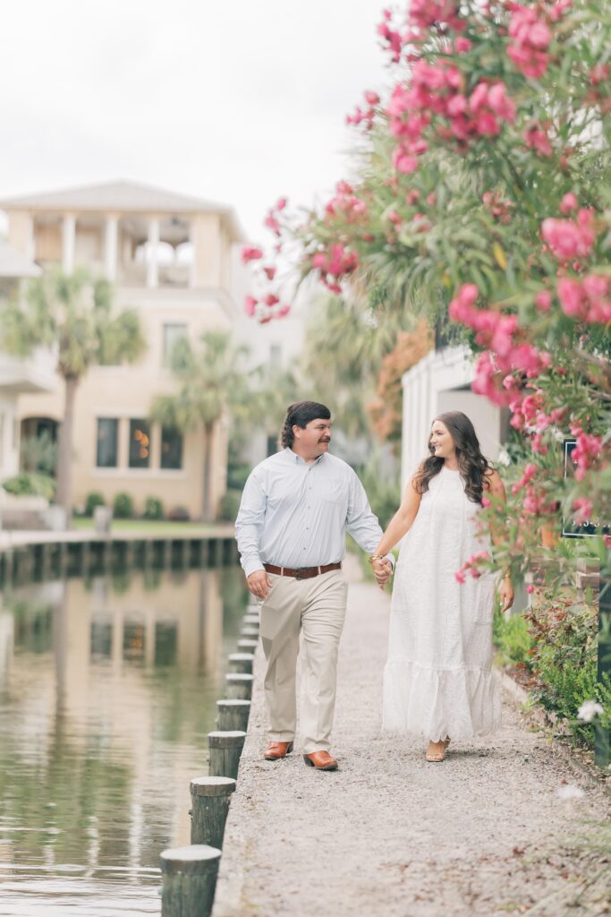 Charleston Wedding Photographer | Engagement Session at Creek at Ion | Couple holding hands walking along the creek with pink flowers in the trees