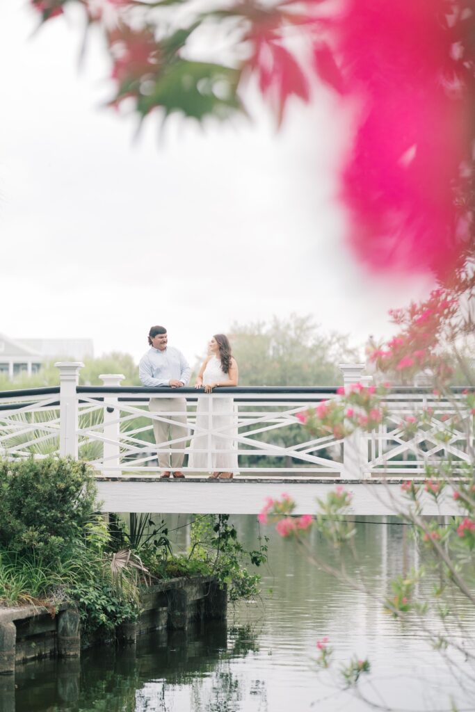 Engagement Photo Locations Columbia SC | Stee Garman Photography Fine-art Wedding Photographer | A couple stands on a white bridge over a calm lake, surrounded by green foliage and vibrant pink flowers. The scene conveys a serene, romantic atmosphere.