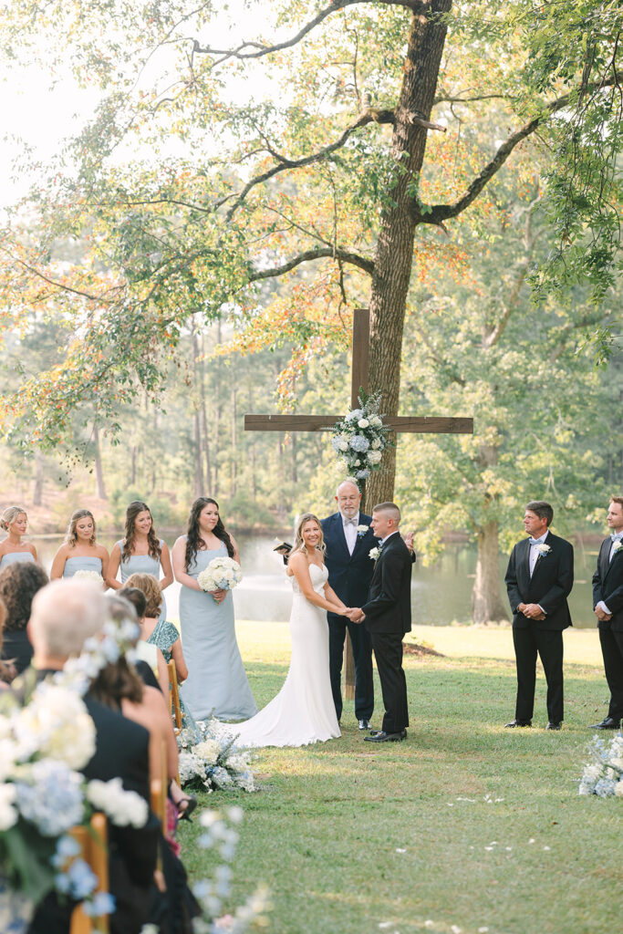 Wedding Photographers in Columbia SC | Stee Garman Photography Fine-art Wedding Photographer | An outdoor wedding with a couple holding hands under a large wooden cross adorned with white flowers. Bridesmaids and groomsmen stand nearby, surrounded by trees and a lake.