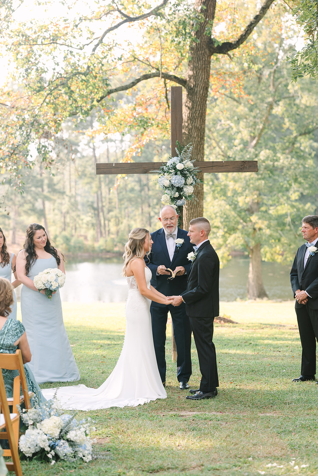 Best Wedding Photographers in Columbia SC | Stee Garman Photography Fine-art Wedding Photographer | Outdoor wedding beside a serene lake with a bride and groom holding hands. An officiant stands under a decorated wooden cross. Bridesmaids and groomsmen watch hands with groom in a black tuxedo under a brick archway and surrounded by greenery