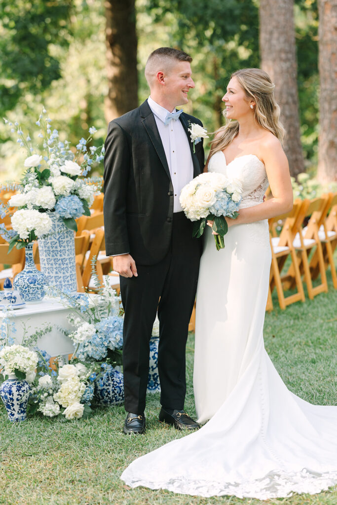 A bride and groom stand smiling outdoors, surrounded by blue and white floral arrangements and ceramic vases.