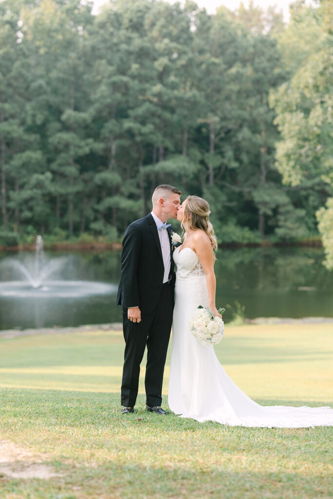Wedding Photographers in Columbia SC | Stee Garman Photography Fine-art Wedding Photographer | Bride and groom share a kiss beside a serene pond with a fountain. Lush green trees in the background; the bride holds a bouquet.