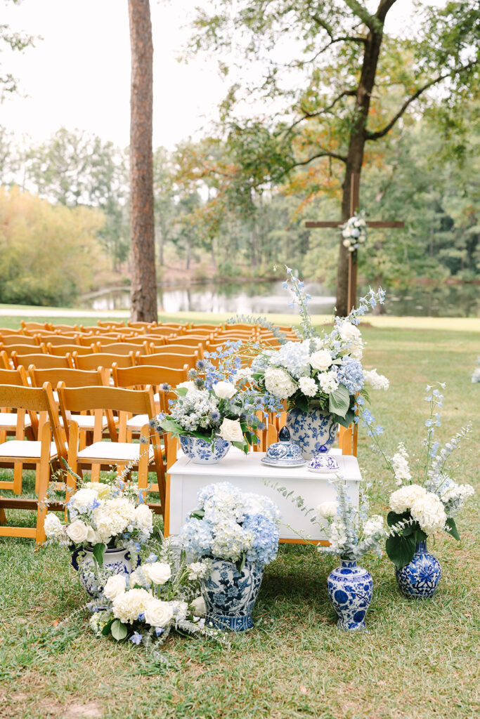 Wedding Photographers in Columbia SC | Stee Garman Photography Fine-art Wedding Photographer | Outdoor wedding setup with wooden chairs on grass. Blue and white floral arrangements in ornate vases. Trees and a lake in the background