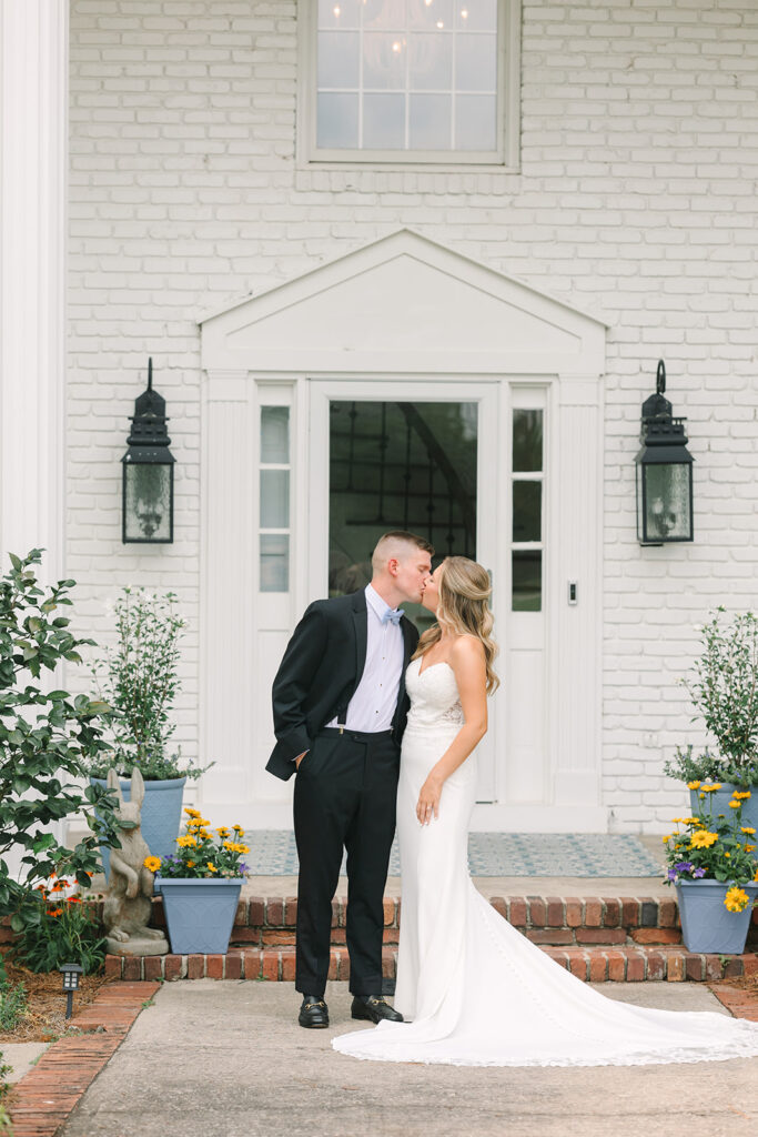Wedding Photographers in Columbia SC | Stee Garman Photography Fine-art Wedding Photographer | A wedding couple shares a kiss in front of a white brick house. The bride wears a strapless white gown, and the groom is in a black suit. Flower pots line the steps, adding color.