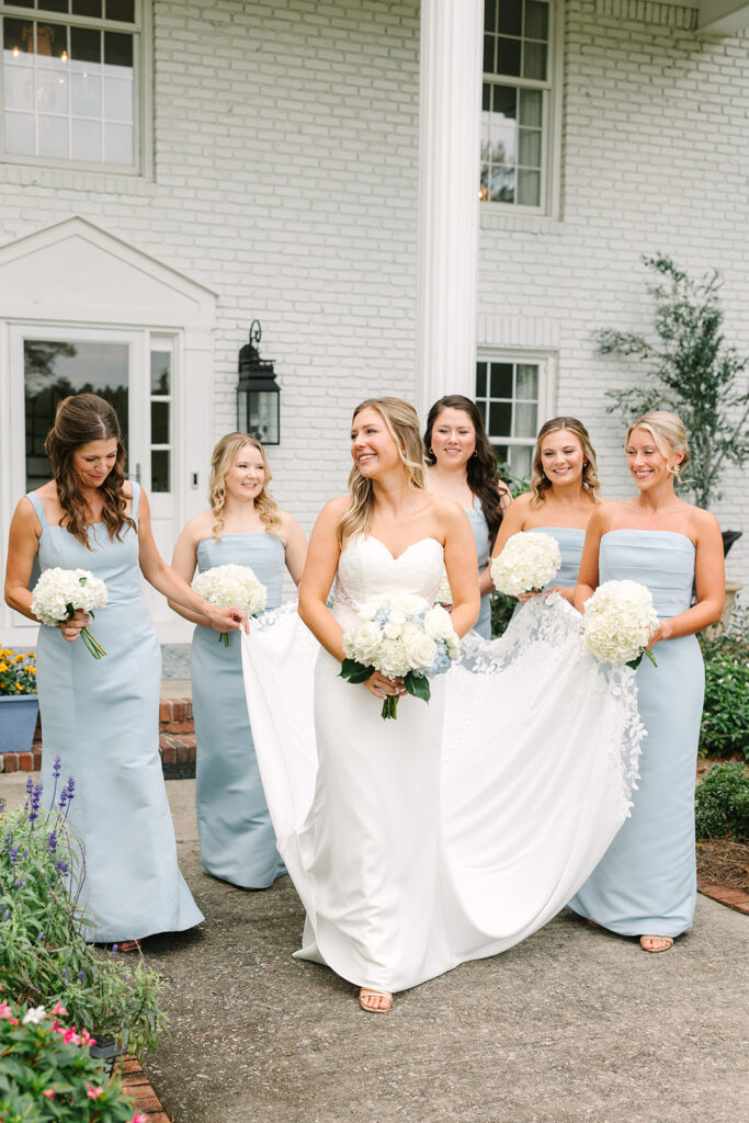 Best Wedding Photographers in Columbia SC | Stee Garman Photography Fine-art Wedding Photographer | Bride in a white gown walking with five bridesmaids wearing light blue dresses, all holding white flower bouquets, smiling in front of a brick building.