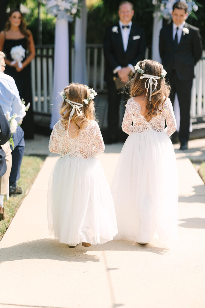 Best Wedding Photographers in Columbia SC | Stee Garman Photography Fine-art Wedding Photographer | Two young flower girls in white lace dresses and floral headbands walk down an aisle. Guests and suited men stand in the background