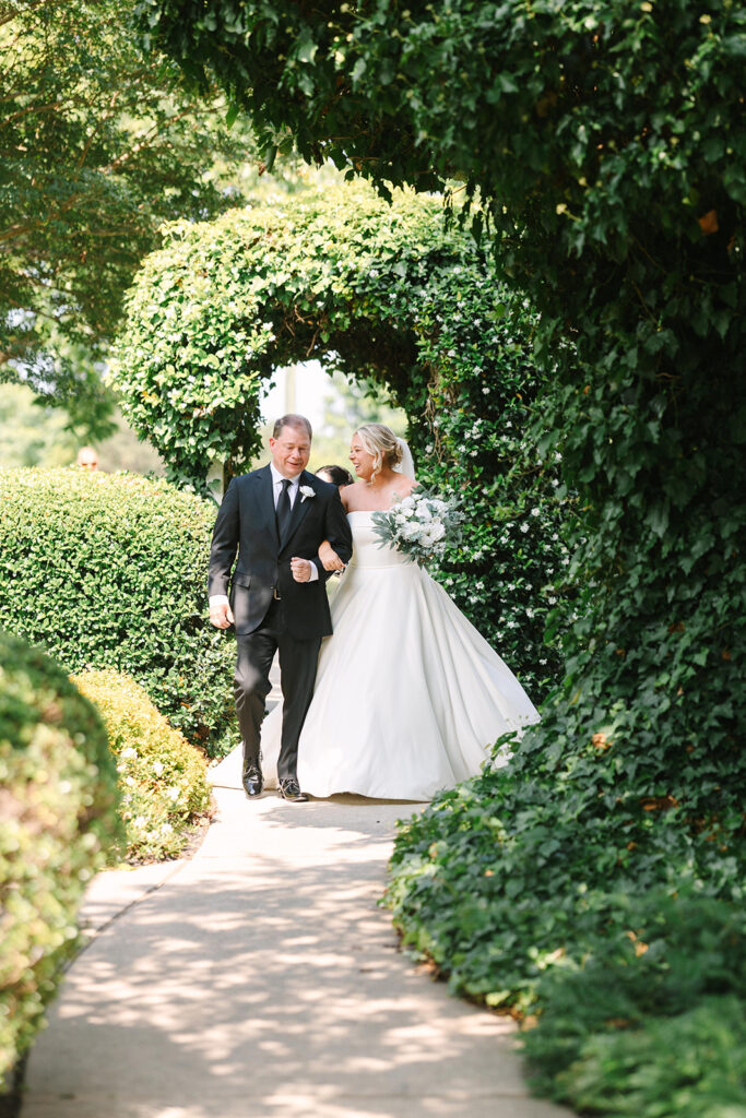 Best Wedding Photographers in Columbia SC | Stee Garman Photography Fine-art Wedding Photographer | A bride in a white gown and her father in a black suit walk arm-in-arm down a garden path framed by lush greenery