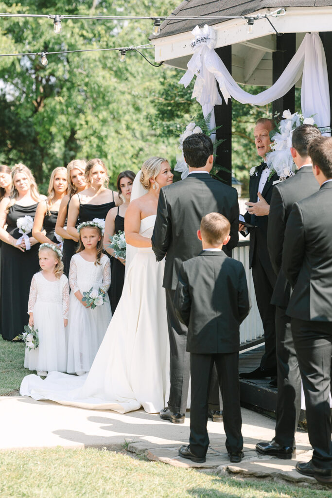 Best Wedding Photographers in Columbia SC | Stee Garman Photography Fine-art Wedding Photographer | A bride and groom stand at an outdoor altar adorned with white drapery. Bridesmaids in black dresses and flower girls in white observe the ceremony.