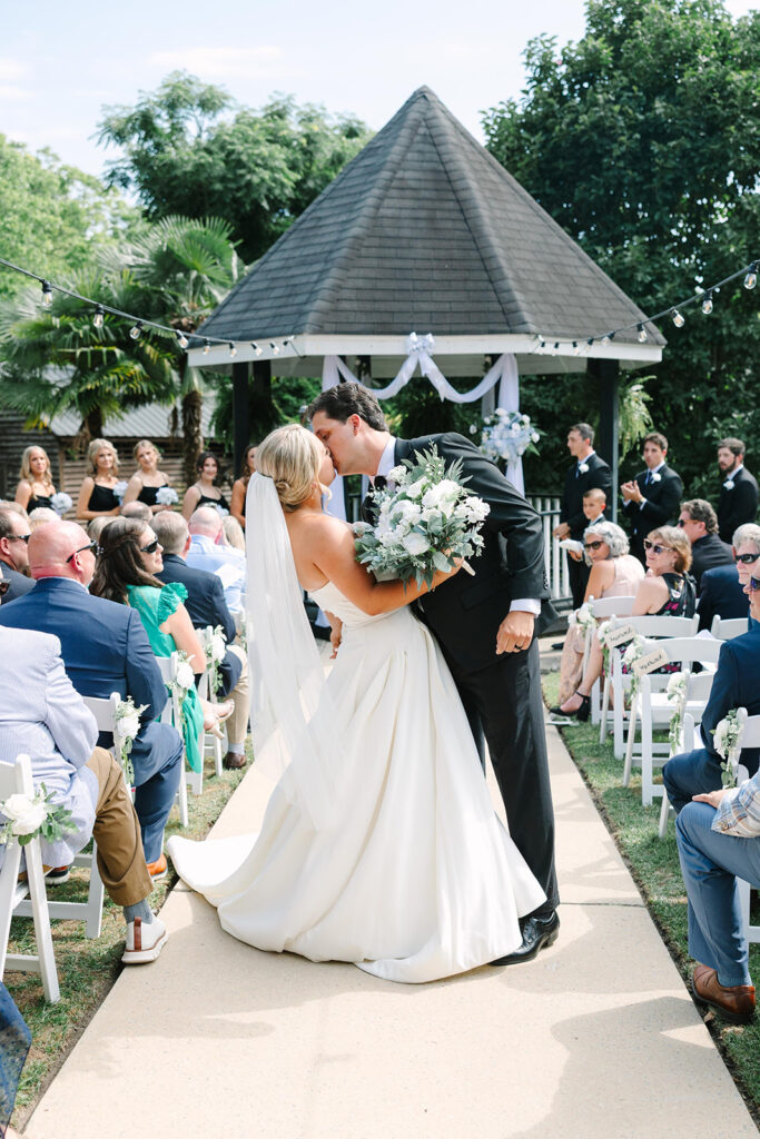 Wedding Photographers in Columbia SC | Stee Garman Photography Fine-art Wedding Photographer | A bride and groom kiss under a gazebo adorned with white flowers in a garden wedding, surrounded by seated guests on a sunny day.
