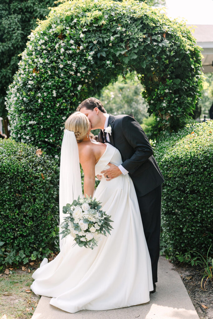 Best Wedding Photographers in Columbia SC | Stee Garman Photography Fine-art Wedding Photographer | A bride and groom share a romantic kiss in front of a lush green archway. The bride holds a bouquet of white flowers, wearing a flowing gown and veil.