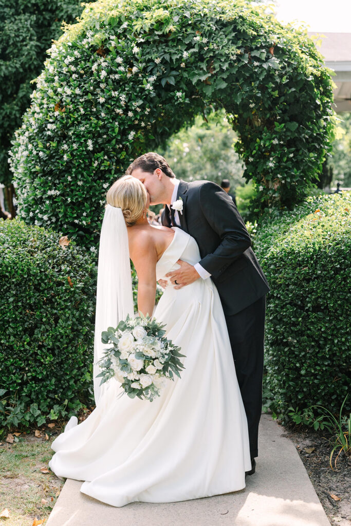 Columbia SC Wedding Photographers | Stee Garman Photography Fine-art Wedding Photographer | Bride and groom share a romantic kiss under a lush green archway. The bride holds a floral bouquet, conveying a joyful, intimate wedding moment.