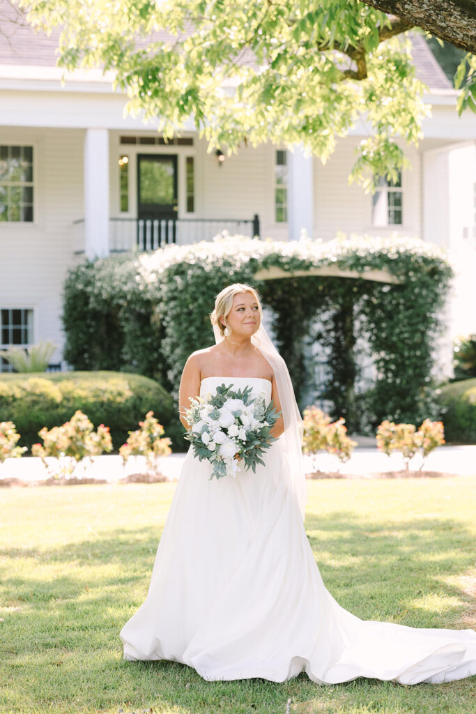 Best Wedding Photographers in Columbia SC | Stee Garman Photography Fine-art Wedding Photographer | Bride in white gown holding a bouquet stands on a sunny lawn, with a charming house and lush greenery in the background