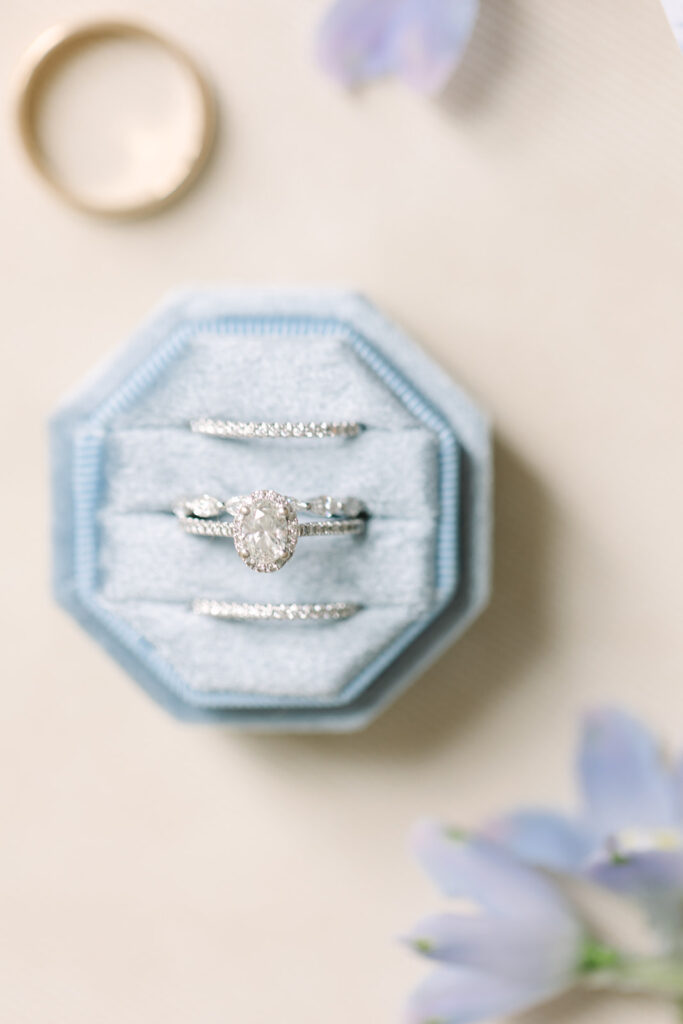 Columbia SC Wedding Photographers | Stee Garman Photography Fine-art Wedding Photographer | A delicate oval diamond ring in a light blue, velvet octagonal box. Beside it, a gold band rests on a soft beige surface with purple flowers nearby.
