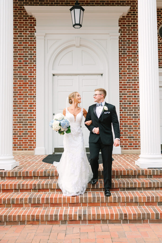 Wedding Photographers in Columbia SC | Stee Garman Photography Fine-art Wedding Photographer | Bride and groom walking arm in arm down brick steps, smiling at each other. Background features white columns and red brick facade.