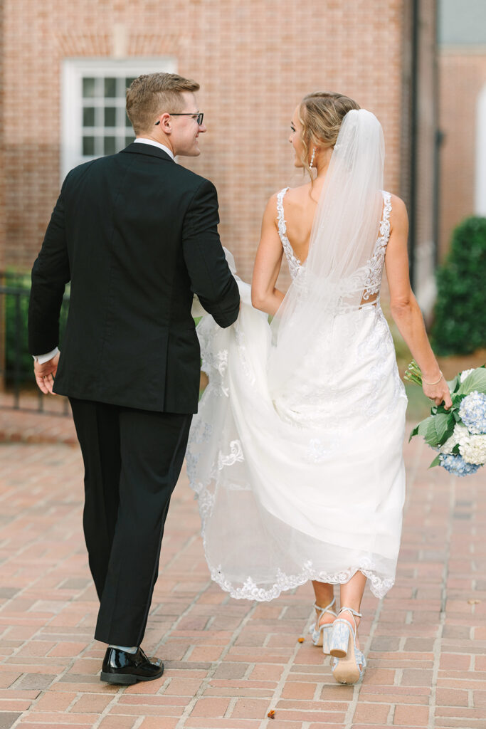 Best Wedding Photographers in Columbia SC | Stee Garman Photography Fine-art Wedding Photographer | Bride in a white lace dress and veil, holding a bouquet, walks hand-in-hand with groom in a black suit. They are smiling on a brick path.