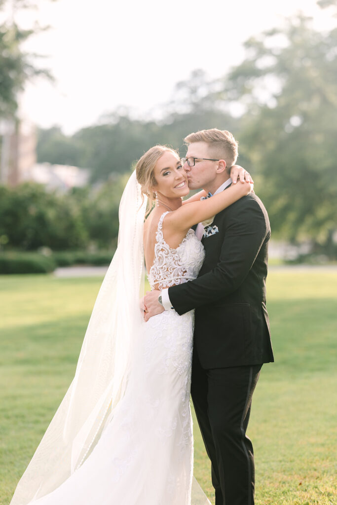 Columbia SC Wedding Photographers | Stee Garman Photography Fine-art Wedding Photographer | A bride in a lace gown and veil hugs a groom in a black suit, smiling joyfully. They stand on a sunny, green lawn with trees in the background.