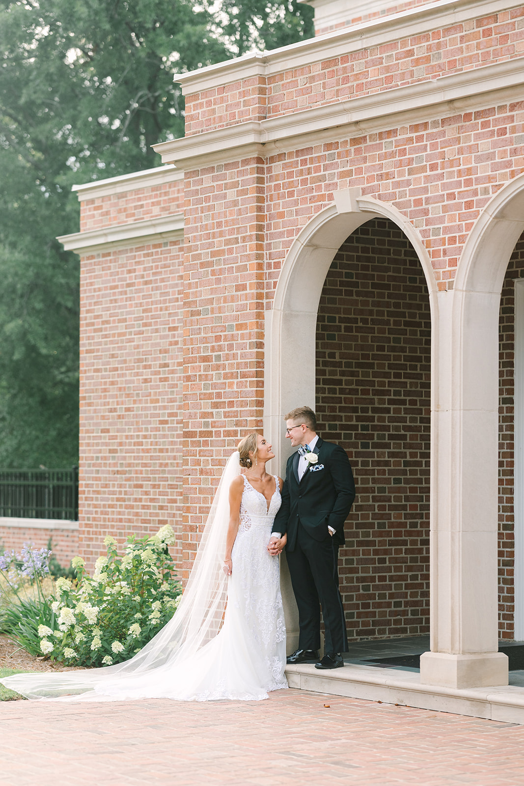 Best Wedding Photographers in Columbia SC | Stee Garman Photography Fine-art Wedding Photographer | Bride in a white lace gown and veil holds hands with groom in a black tuxedo under a brick archway and surrounded by greenery