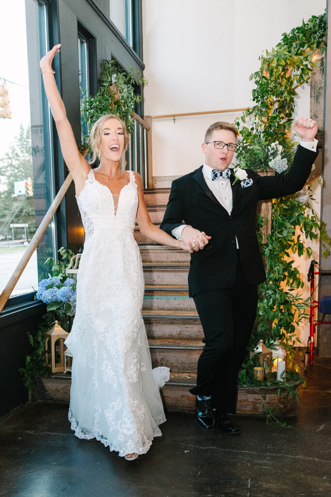 Columbia SC Wedding Photographers | Stee Garman Photography Fine-art Wedding Photographer | A joyful couple enters a wedding venue. The bride, in a lace gown, raises her arm in celebration. The groom, in a tuxedo, follows suit. Lush greenery decorates the wooden stairs behind them.
