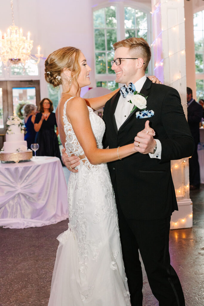 Columbia SC Wedding Photographers | Stee Garman Photography Fine-art Wedding Photographer | A bride in a lace gown and a groom in a tuxedo share a joyful dance at a wedding reception. A cake is visible in the background under soft chandelier light.
