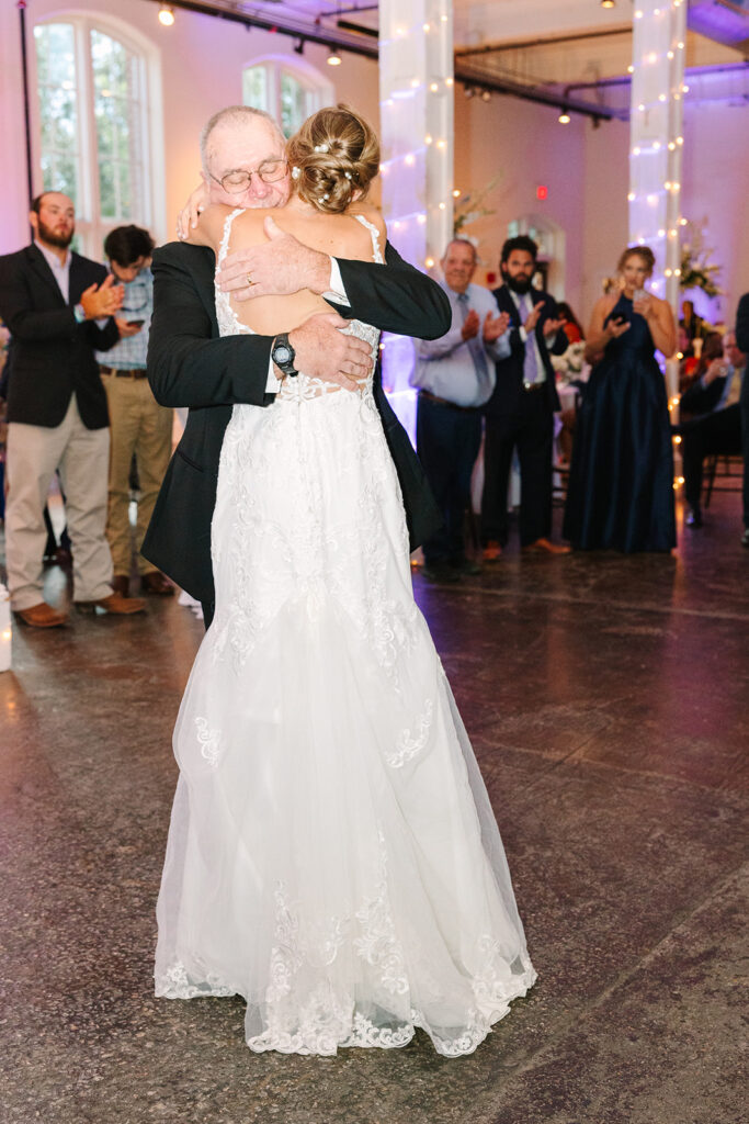 Columbia SC Wedding Photographers | Stee Garman Photography Fine-art Wedding Photographer | A bride in a lacy white gown hugs her father in a suit during a wedding dance. Guests in elegant attire clap warmly in the softly lit venue.