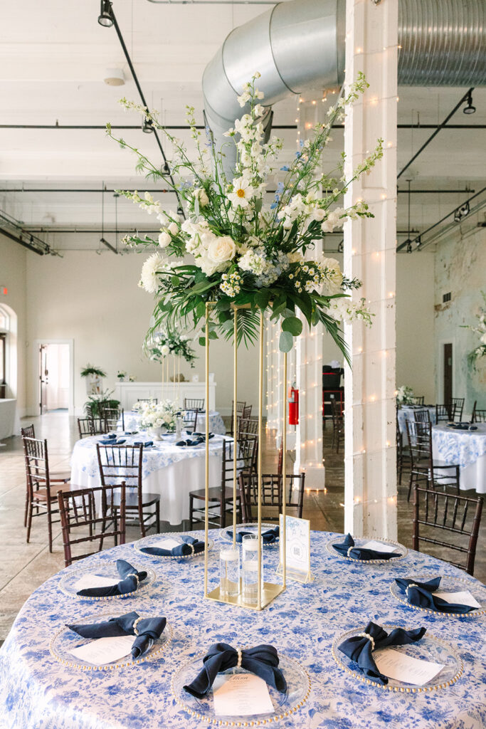 A tall floral centerpiece rises above blue-and-white linens inside an industrial reception space, ideal for modern wedding venues in Columbia SC.