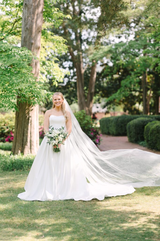 Outdoor Wedding Venues Columbia SC | Stee Garman Photography Fine-art Wedding Photographer | A bride in a flowing white gown and veil smiles while holding a bouquet in a sunlit garden. Lush greenery and blooming flowers create a serene backdrop.