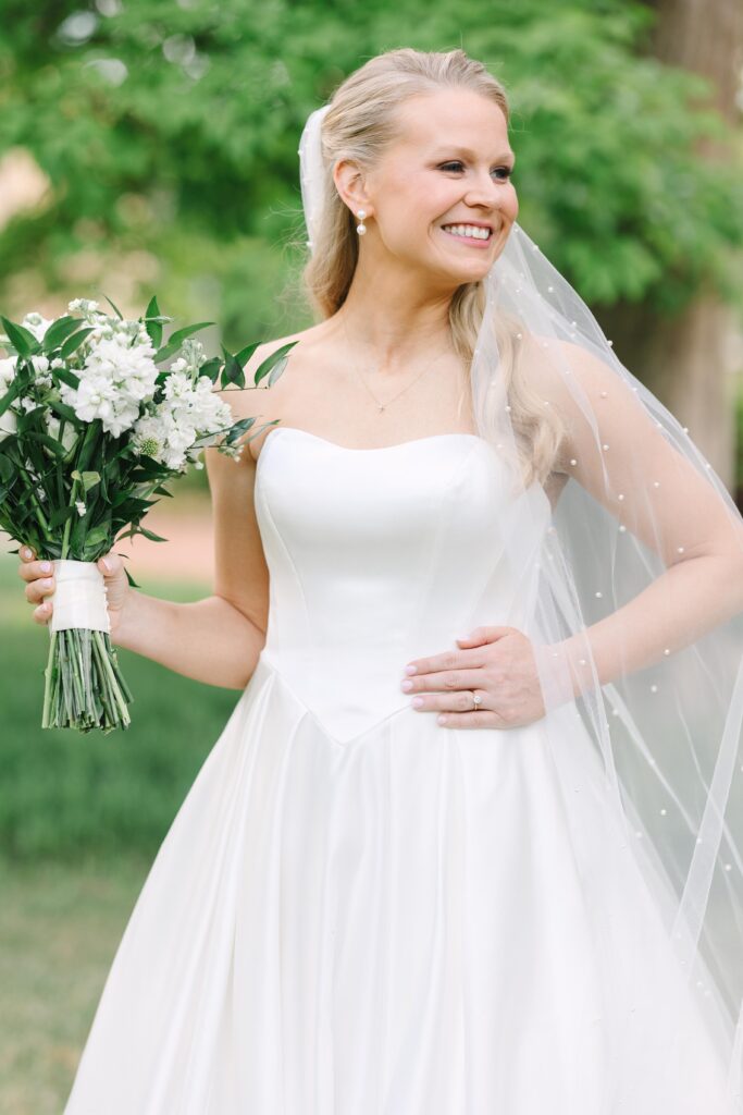 Columbia SC Wedding Photographers | Stee Garman Photography Fine-art Wedding Photographer | A smiling bride in a white gown and veil holds a bouquet of white flowers. She stands outdoors with green trees in the background.