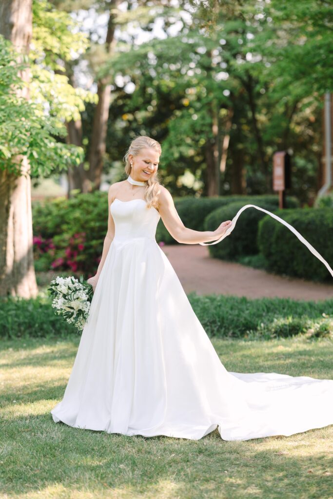 Bridal Portraits Photographer Columbia SC | Stee Garman Photography Fine-art Wedding Photographer | A smiling bride in a flowing white gown with a long train stands in a lush garden holding a bouquet. Sunlight filters through green trees, creating a serene and joyful atmosphere.