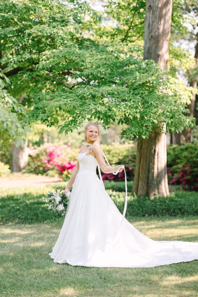 South Carolina Wedding Photographers | Stee Garman Photography Fine-art Wedding Photographer | A bride in a flowing white gown smiles under lush green trees, holding a bouquet. Bright, colorful flowers and sunlight create a joyful, serene scene.