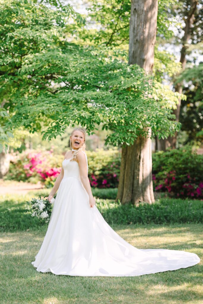 Wedding Photographers Columbia SC | Stee Garman Photography Fine-art Wedding Photographer | Bride in elegant white gown joyfully laughing in a sunlit garden, holding a bouquet. Lush greenery and vibrant pink flowers create a serene backdrop.