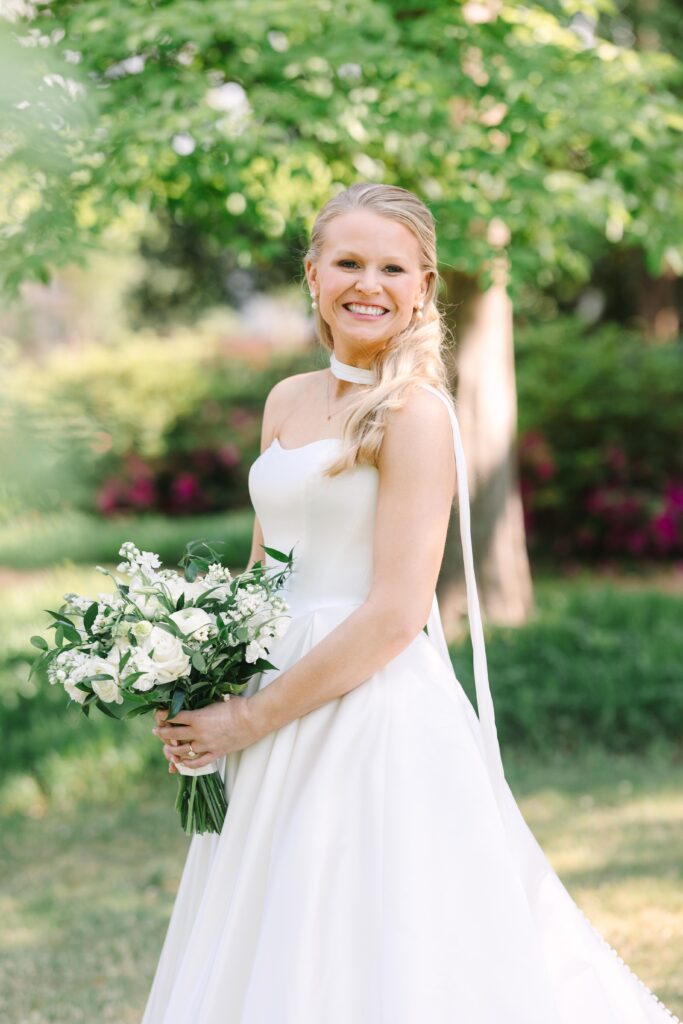 Bridal Portraits Photographer Columbia SC | Stee Garman Photography Fine-art Wedding Photographer | Bride in white gown holding a bouquet of white flowers, smiling in a sunlit garden. Green foliage and vibrant flower beds create a joyful, serene backdrop.