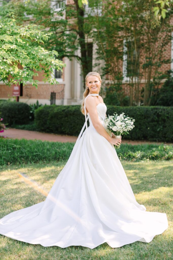 Wedding Photographers Columbia SC | Stee Garman Photography Fine-art Wedding Photographer | A bride in a flowing white gown stands on a sunlit grassy lawn, holding a bouquet of white flowers. She smiles warmly, exuding joy and elegance.