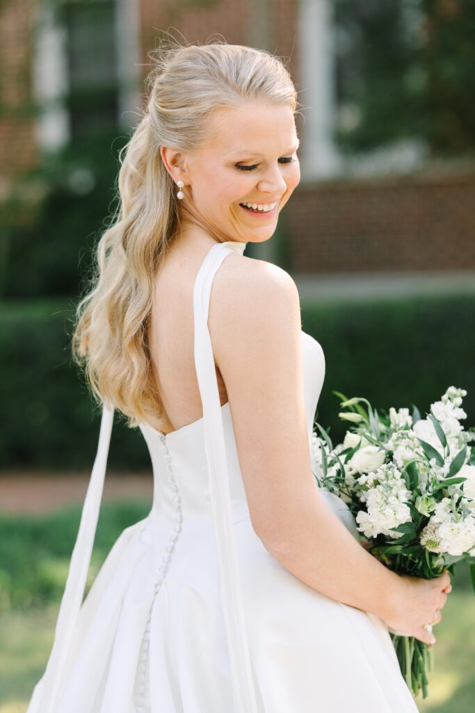 Bridal Portraits Photographer Columbia SC | Stee Garman Photography Fine-art Wedding Photographer | Bride smiling while holding a bouquet of white flowers. She has long, wavy hair and wears a white dress with a buttoned back. The background is blurred greenery.