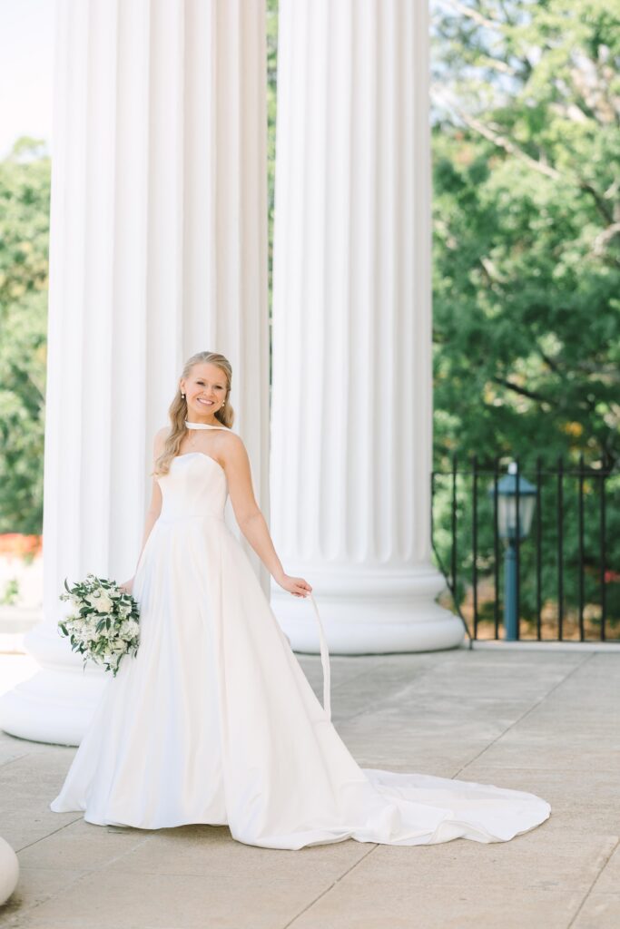 Wedding Photographers Columbia SC | Stee Garman Photography Fine-art Wedding Photographer | Bride in a white gown stands joyfully in front of large white columns, holding a bouquet. Green trees provide a natural, serene backdrop.