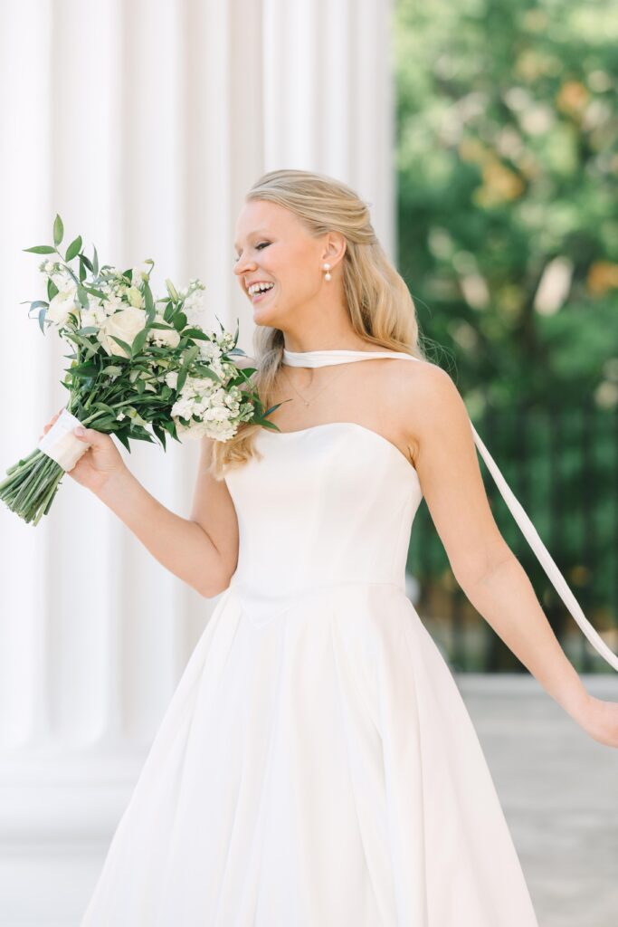 Wedding Photographers Columbia SC | Stee Garman Photography Fine-art Wedding Photographer | A joyful bride in a strapless white gown holds a lush bouquet of green and white flowers. She stands outside, smiling, with sunlit greenery in the background.