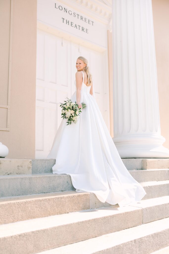 Wedding Photographers Columbia SC | Stee Garman Photography Fine-art Wedding Photographer | A bride in a flowing white gown smiles while holding a bouquet on stone steps. The scene is bright and elegant, set in front of a building with large columns.