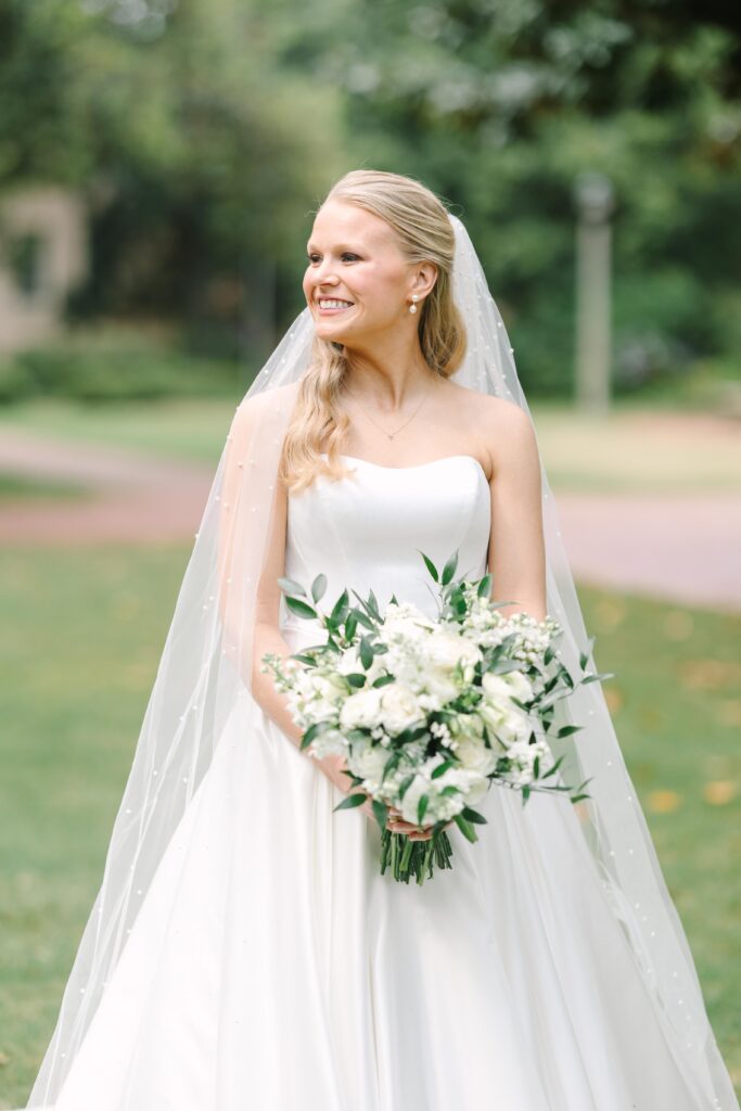 Wedding Photographers Columbia SC | Stee Garman Photography Fine-art Wedding Photographer | A smiling bride in a white gown and pearl-studded veil holds a lush bouquet of white flowers and greenery, standing in a sunlit, leafy garden.