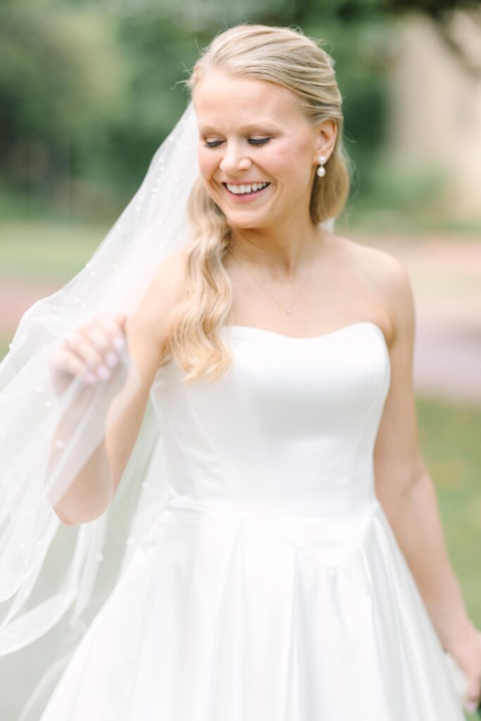 Bridal Portraits Photographer Columbia SC | Stee Garman Photography Fine-art Wedding Photographer | A smiling bride in a strapless white gown and veil, with wavy blonde hair, stands outdoors. The background is softly blurred with greenery, conveying joy.