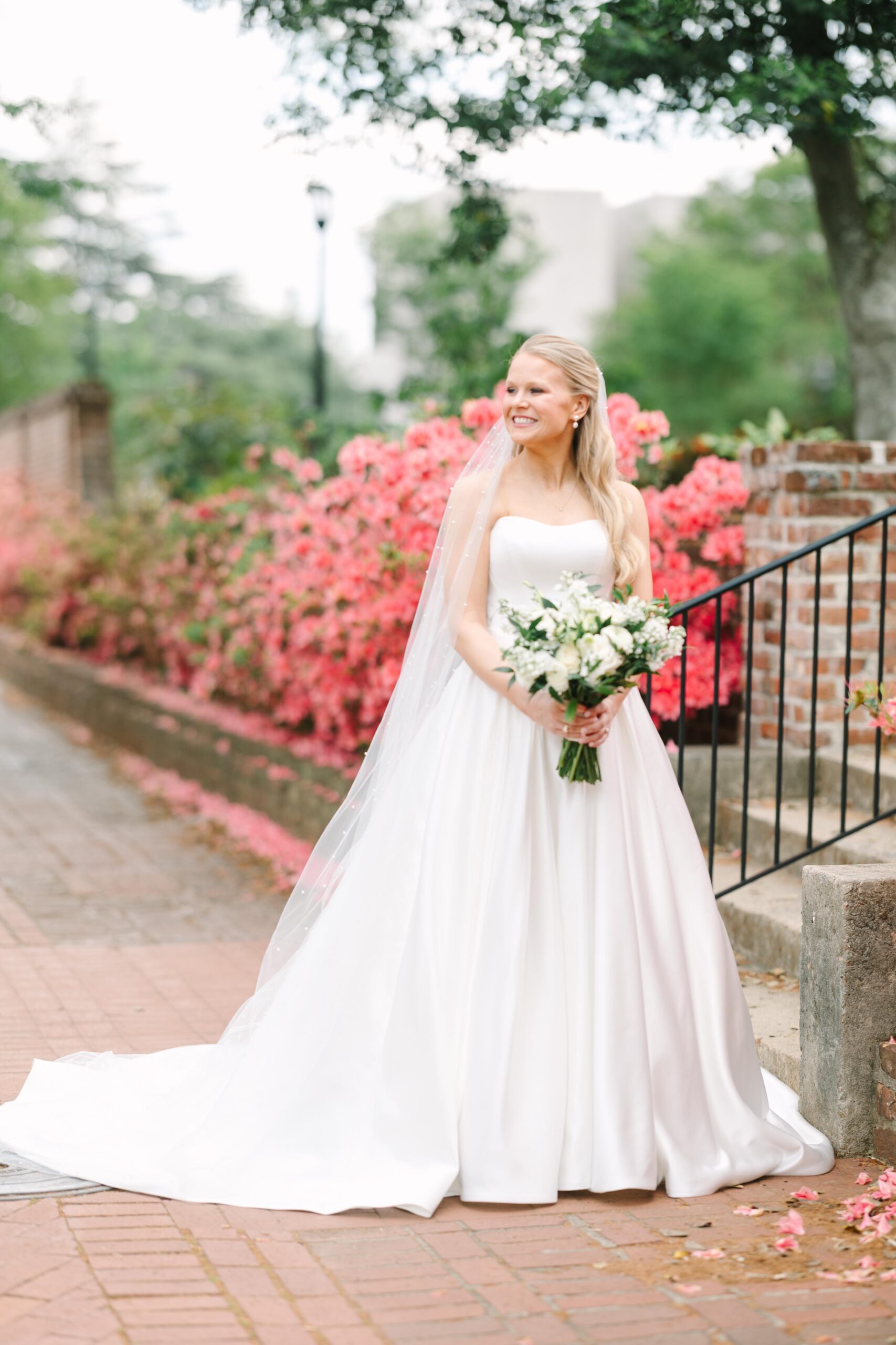 Bridal Portraits Photographer Columbia SC | Stee Garman Photography Fine-art Wedding Photographer | Bride in a flowing white gown and veil holds a bouquet, smiling joyfully. She's standing beside vibrant pink flowers and a brick wall. Lush greenery in the background.