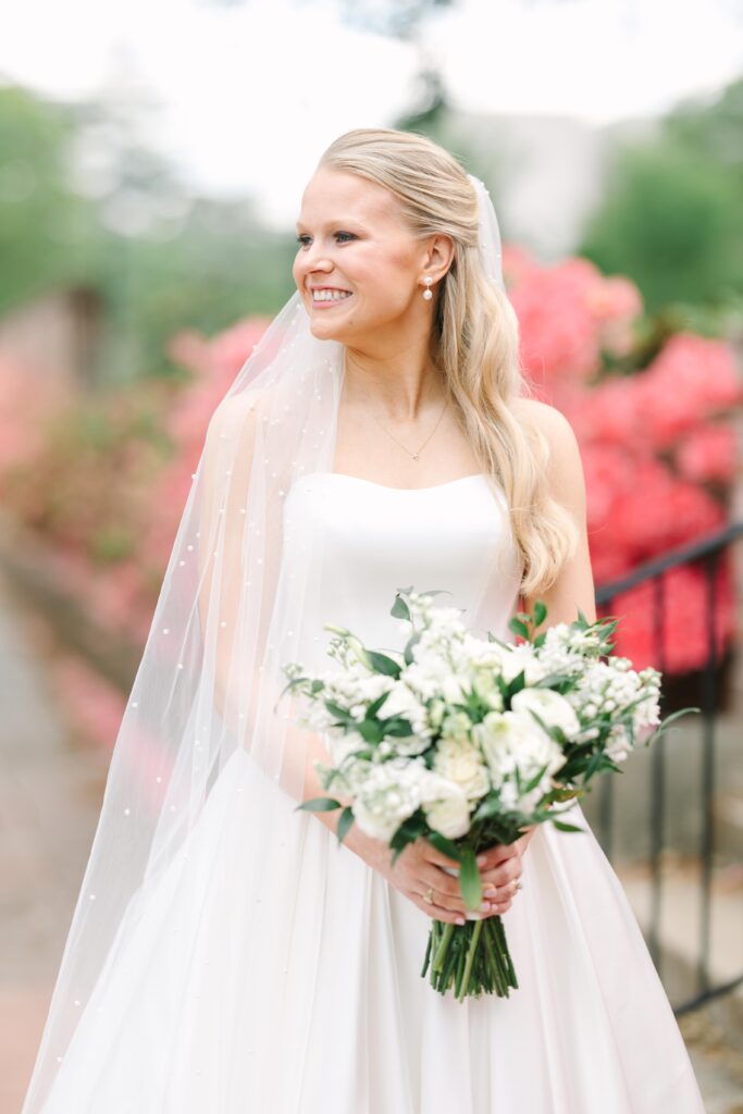 Wedding Photographers Columbia SC | Stee Garman Photography Fine-art Wedding Photographer | A smiling bride in a white gown and veil holds a bouquet of white flowers. She's outdoors, with vibrant pink flowers in the blurred background, conveying joy.