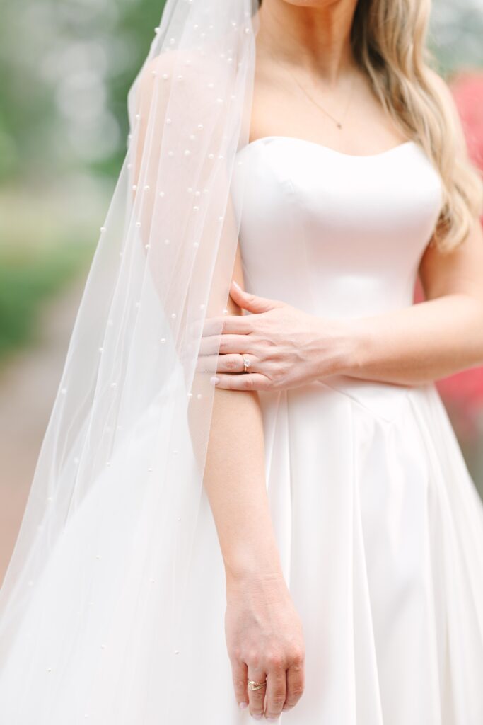 Wedding Photographers Columbia SC | Stee Garman Photography Fine-art Wedding Photographer | Bride in an elegant white dress with pearl-embellished veil, embracing her arm. Soft background of greenery and pink flowers.
