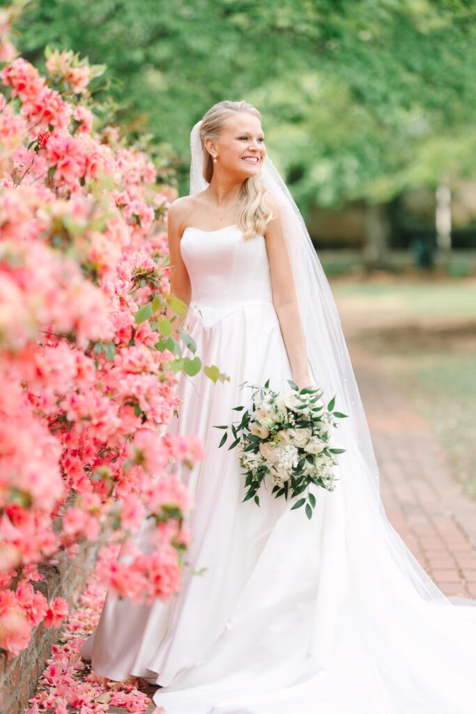 Columbia SC Wedding Photographers | Stee Garman Photography Fine-art Wedding Photographer | A joyful bride in a strapless white gown and veil stands beside vibrant pink flowers, holding a lush bouquet, set against a serene garden backdrop.