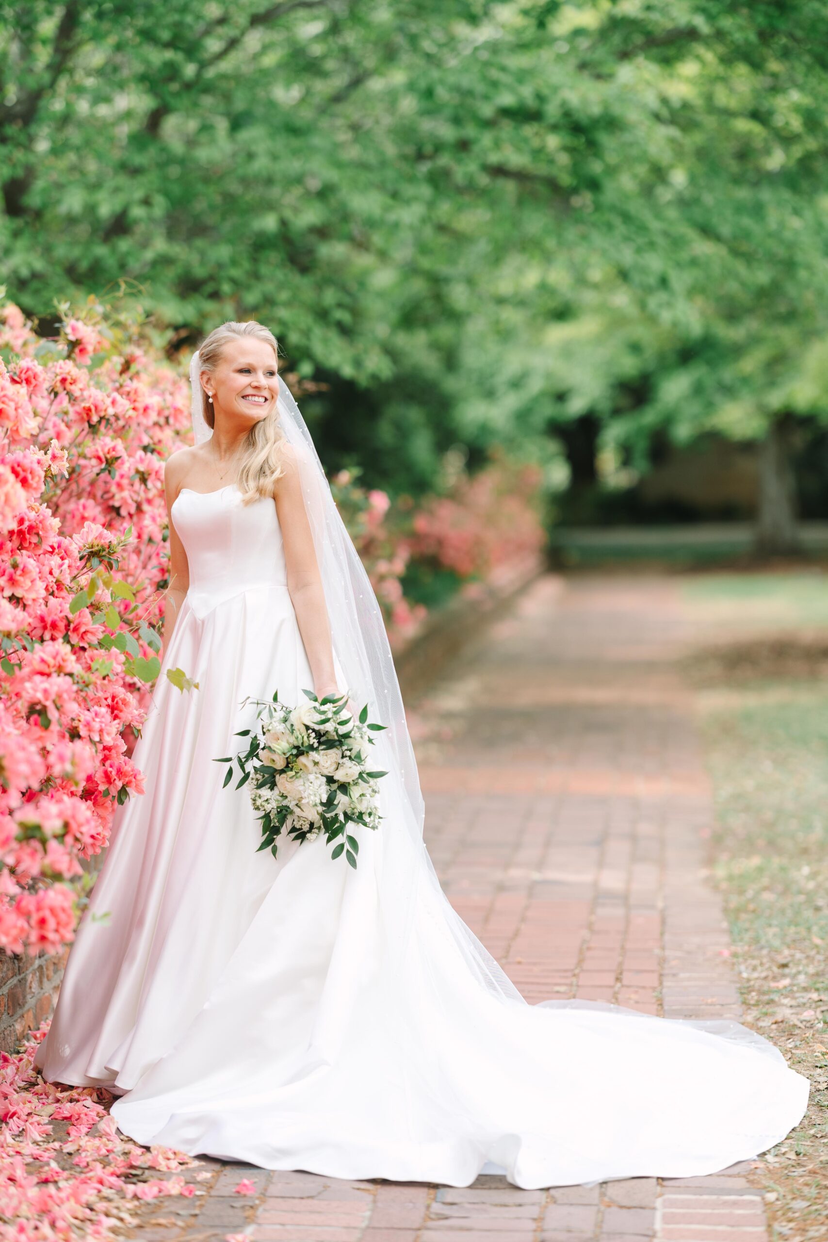Columbia SC Wedding Photographers | Stee Garman Photography Fine-art Wedding Photographer | A joyful bride in a strapless white gown and veil stands beside vibrant pink flowers, holding a lush bouquet, set against a serene garden backdrop.