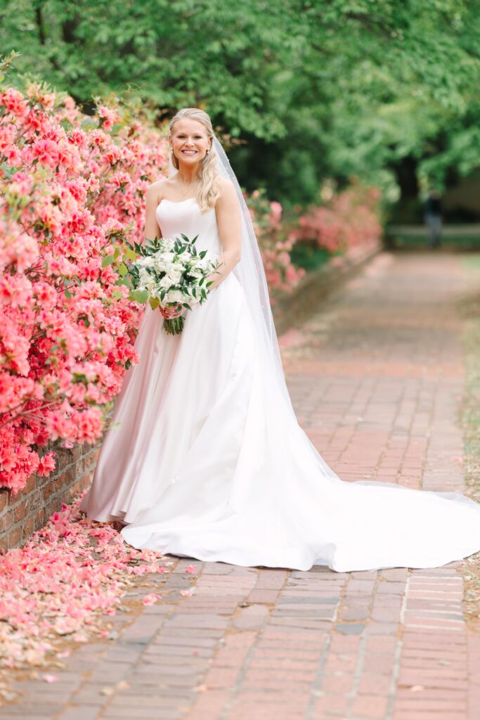 Wedding Photographers Columbia SC | Stee Garman Photography Fine-art Wedding Photographer | Bride in a flowing white gown stands smiling by vibrant pink flowers on a brick path. She holds a bouquet, conveying joy and serenity.