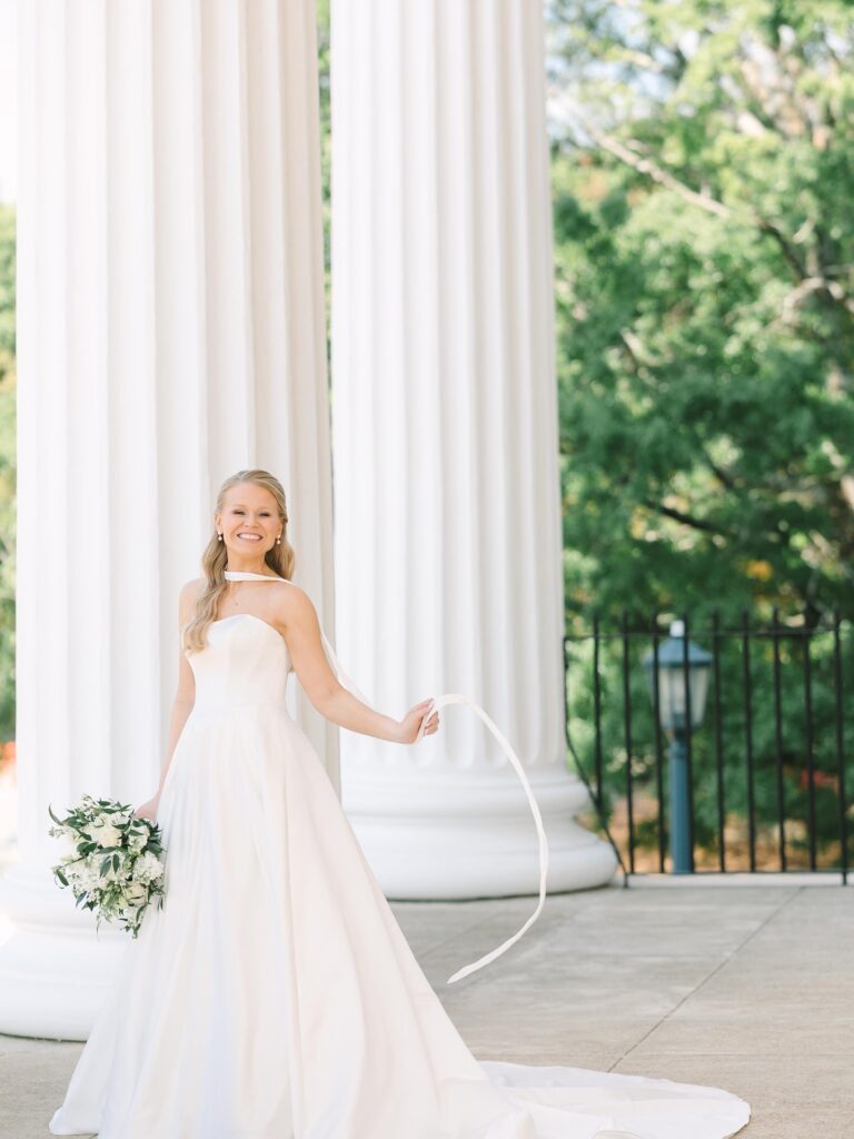Columbia SC Wedding Photographers | Stee Garman Photography Fine-art Wedding Photographer | A smiling bride in a strapless white gown stands near large white columns, holding a bouquet.