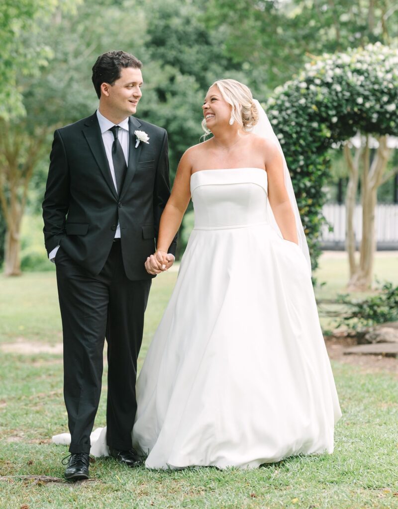 Columbia SC Wedding Photographers | Stee Garman Photography Fine-art Wedding Photographer | A joyful bride and groom walk hand in hand outdoors. She wears a flowing white gown, and he dons a black suit. Lush greenery surrounds them